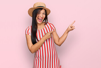 Young hispanic woman wearing summer hat smiling and looking at the camera pointing with two hands...