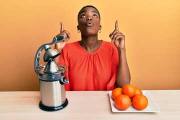 Young african american woman sitting on the table using juicer amazed and surprised looking up and...