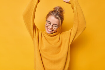 Positive carefree woman raises arms up dances carefree from joy wears round transparent glasses and jumper smiles gladfully keeps eyes closed isolated over vivid yellow background rejoices success