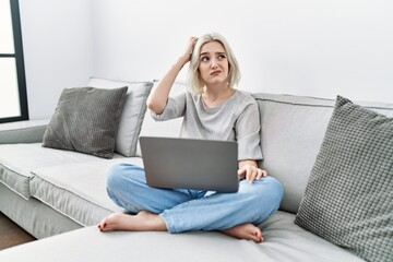 Young caucasian woman using laptop at home sitting on the sofa confuse and wonder about question....
