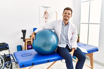 Young hispanic man wearing physiotherapist uniform holding fit ball at clinic