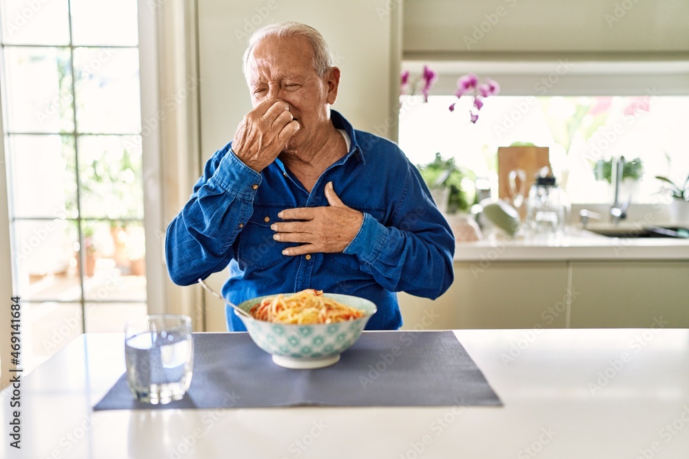 Canvas Prints Senior man with grey hair eating pasta spaghetti at home smelling something stinky and disgusting, intolerable smell, holding breath with fingers on nose. bad smell