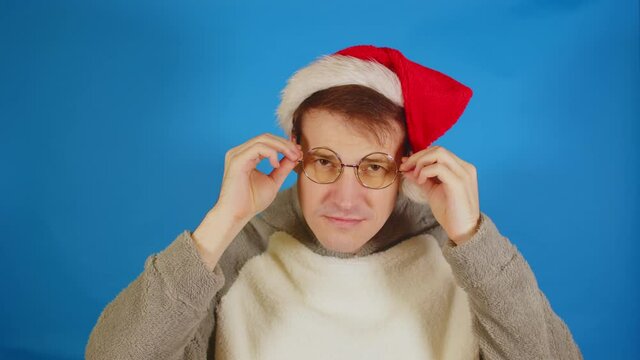 Man in santa hat puts on glasses, standing on blue background. Male in christmas hat and eyeglasses looking at camera.
