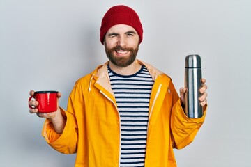 Caucasian man with beard holding cup of coffee and thermo winking looking at the camera with sexy...