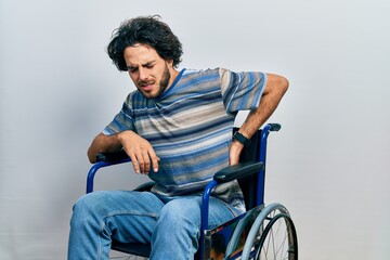Handsome hispanic man sitting on wheelchair suffering of backache, touching back with hand,...