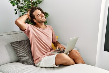 Young hispanic man sitting on the sofa at home using laptop smiling confident touching hair with...