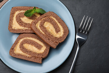 Tasty chocolate cake roll with cream and mint on black table, flat lay