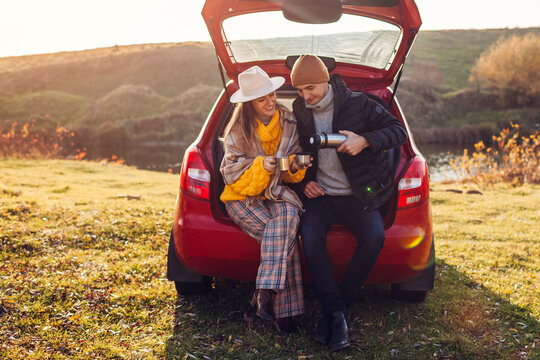 Loving couple relaxing in car trunk by autumn lake drinking tea from vacuum flask. Man and woman travel by auto.
