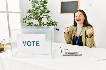 Young brunette woman sitting at election table with voting ballot very happy and excited doing...