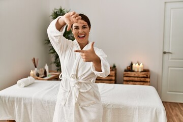 Fototapeta na wymiar Young beautiful hispanic woman wearing bathrobe at wellness spa smiling making frame with hands and fingers with happy face. creativity and photography concept.