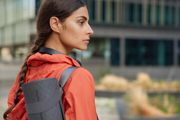 Back view of sporty woman with pigtail dressed in anorak keeps healthy has regular training uses sport accessories looks away concentrated into distance poses outdoors against blurred background