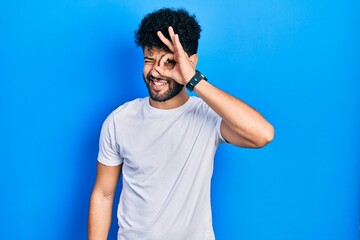 Fototapeta premium Young arab man with beard wearing casual white t shirt doing ok gesture with hand smiling, eye looking through fingers with happy face.