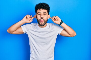 Young arab man with beard wearing casual white t shirt smiling pulling ears with fingers, funny...