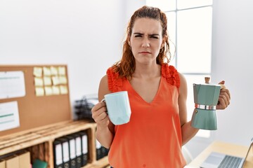 Young redhead woman holding french coffee maker at the office clueless and confused expression....