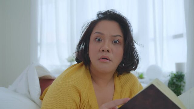 Asian woman oversize big chubby girl lying in bed reading a book in the bedroom showing a panic look at the camera. Leisure and hobbies on holiday.