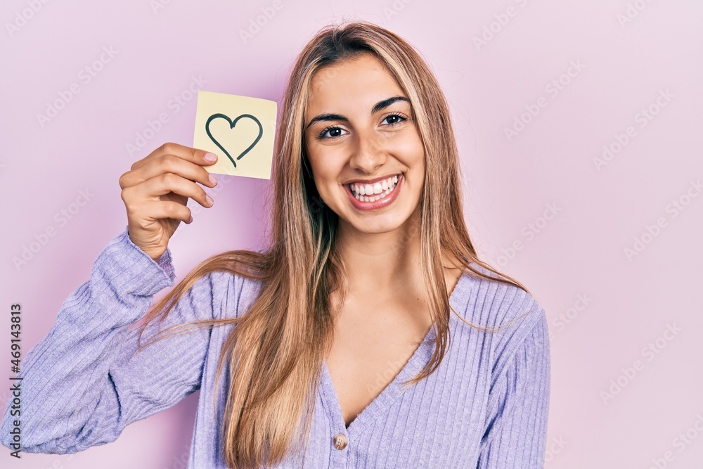 Canvas Prints Beautiful hispanic woman holding heart reminder looking positive and happy standing and smiling with a confident smile showing teeth