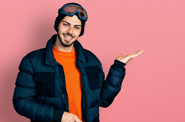 Young hispanic man with beard wearing snow wear and sky glasses smiling cheerful presenting and pointing with palm of hand looking at the camera.