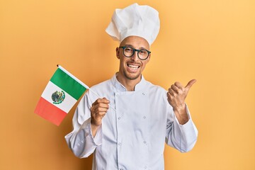 Bald man with beard wearing professional cook apron holding mexico flag pointing thumb up to the...