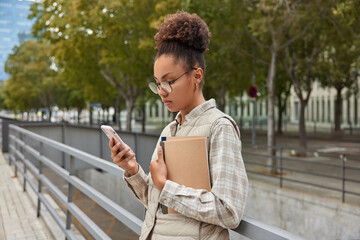 Fototapeta premium Intelligent female student concentrated at smartphone screen learns schedule for next week holds textbook wears round spectacles and casual clothes poses at city against trees uses modern technologies