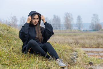 Naklejka na ściany i meble A beautiful girl with long hair in black clothes sits on the grass on the outskirts of the city. She puts a hood over her head and looks away