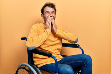Handsome man with beard sitting on wheelchair laughing and embarrassed giggle covering mouth with...