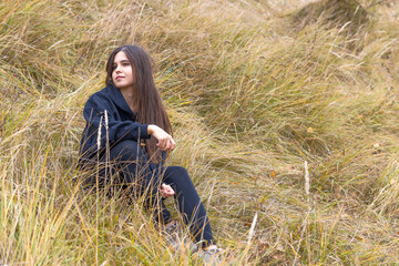 Naklejka na ściany i meble A beautiful girl with long hair in black clothes sits on a hillside among the thick autumn grass