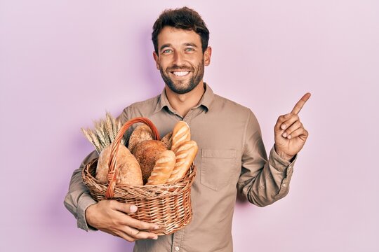 Handsome man with beard holding wicker basket with bread smiling happy pointing with hand and finger to the side