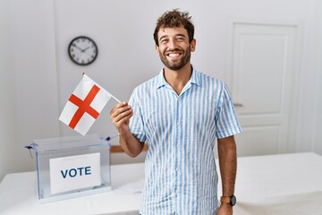 Young handsome man at political campaign election holding england flag looking positive and happy standing and smiling with a confident smile showing teeth