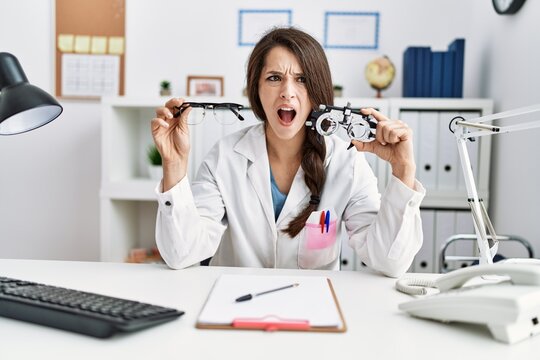 Young doctor woman holding optometry glasses and normal glasses angry and mad screaming frustrated and furious, shouting with anger. rage and aggressive concept.