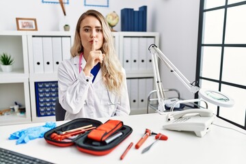Young beautiful doctor woman with reflex hammer and medical instruments asking to be quiet with...