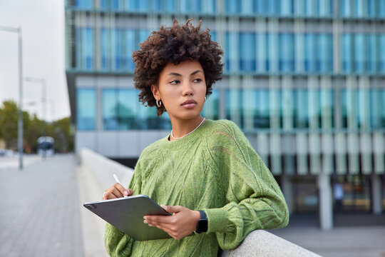 Photo of thoughtful woman with cury hair uses stylus pen to drw graphics on digital tablet works with inspiration wears casual jumper poses against modern city building creats content for website