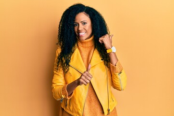 Middle age african american woman wearing wool winter sweater and leather jacket pointing to the back behind with hand and thumbs up, smiling confident