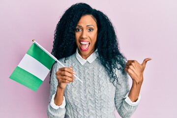 Middle age african american woman holding nigeria flag pointing thumb up to the side smiling happy with open mouth