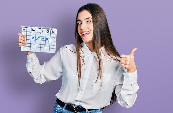 Young brunette teenager holding travel calendar smiling happy and positive, thumb up doing excellent and approval sign
