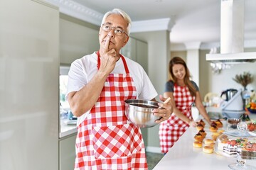 Fototapeta na wymiar Middle age hispanic couple wearing apron cooking homemade pastry serious face thinking about question with hand on chin, thoughtful about confusing idea