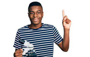 Young african american man holding microscope smiling with an idea or question pointing finger with happy face, number one