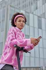 Vertical shot of young woman uses scooter as means of transport sends message to friend leads active lifestyle enjoys day off wears sweater and headband concentrate away. Eco transportation.