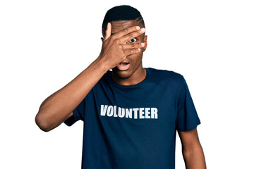 Fototapeta premium Young african american man wearing volunteer t shirt peeking in shock covering face and eyes with hand, looking through fingers with embarrassed expression.