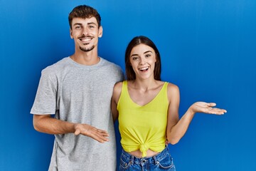 Fototapeta premium Young hispanic couple standing together over blue background pointing aside with hands open palms showing copy space, presenting advertisement smiling excited happy