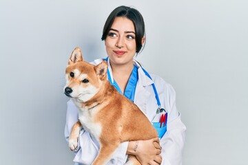 Beautiful hispanic veterinarian woman holding dog smiling looking to the side and staring away...