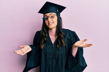 Young hispanic woman wearing graduation cap and ceremony robe clueless and confused expression with...