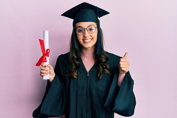 Young hispanic woman wearing graduation uniform holding diploma pointing thumb up to the side smiling happy with open mouth - obrazy, fototapety, plakaty