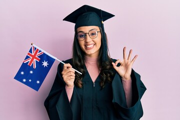 Young hispanic woman wearing graduation uniform holding australia flag doing ok sign with fingers, smiling friendly gesturing excellent symbol