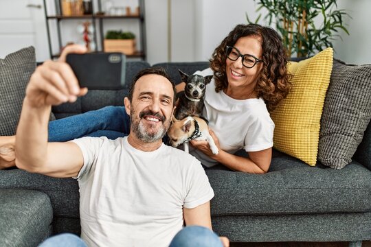 Middle age hispanic couple smiling happy and making selfie by the smartphone. Sitting on the sofa with dogs at home.