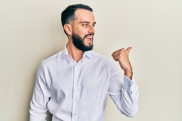 Young man with beard wearing business white shirt pointing thumb up to the side smiling happy with open mouth