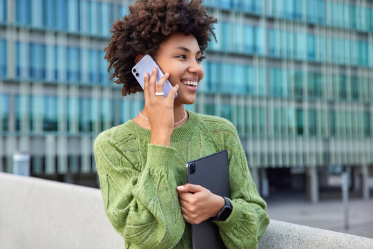 Photo of attractive cheerful woman uses modern technologies modern smartphone for calling wears casual clothes poses outside against blurred city background discusses interesting topic with friend - Powered by Adobe