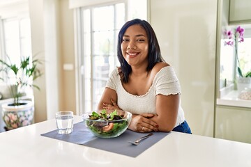 Obraz na płótnie Canvas Young hispanic woman eating healthy salad at home happy face smiling with crossed arms looking at the camera. positive person.