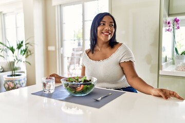 Obraz na płótnie Canvas Young hispanic woman eating healthy salad at home looking away to side with smile on face, natural expression. laughing confident.