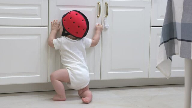 Curious toddler wearing safety helmet exploring kitchen cupboards, child trying to open the kitchen cabinet door with baby safety lock. High quality 4k footage