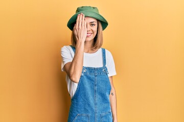 Young caucasian blonde woman wearing denim jumpsuit and hat with 90s style covering one eye with...